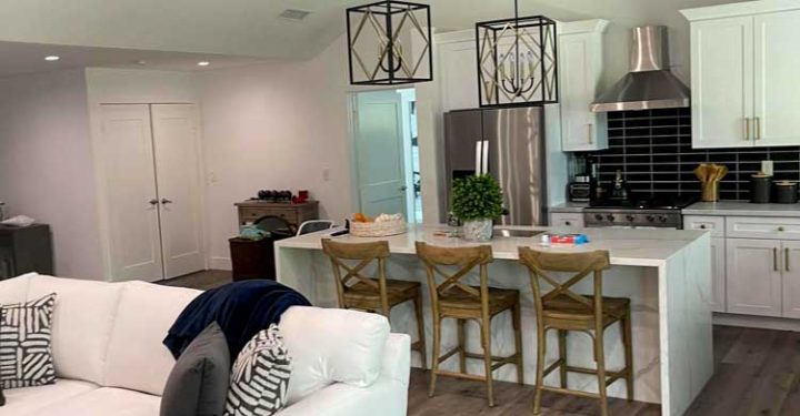 a living room couch, kitchen area in the background, white kitchen cabinet, kitchen island with three wooden chairs