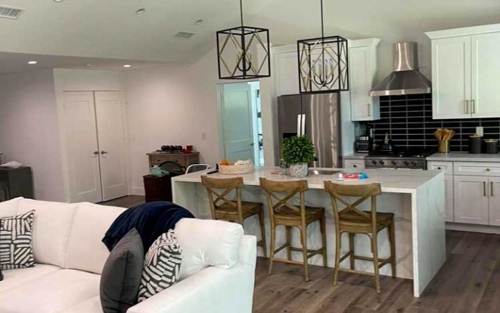 a living room couch, kitchen area in the background, white kitchen cabinet, kitchen island with three wooden chairs